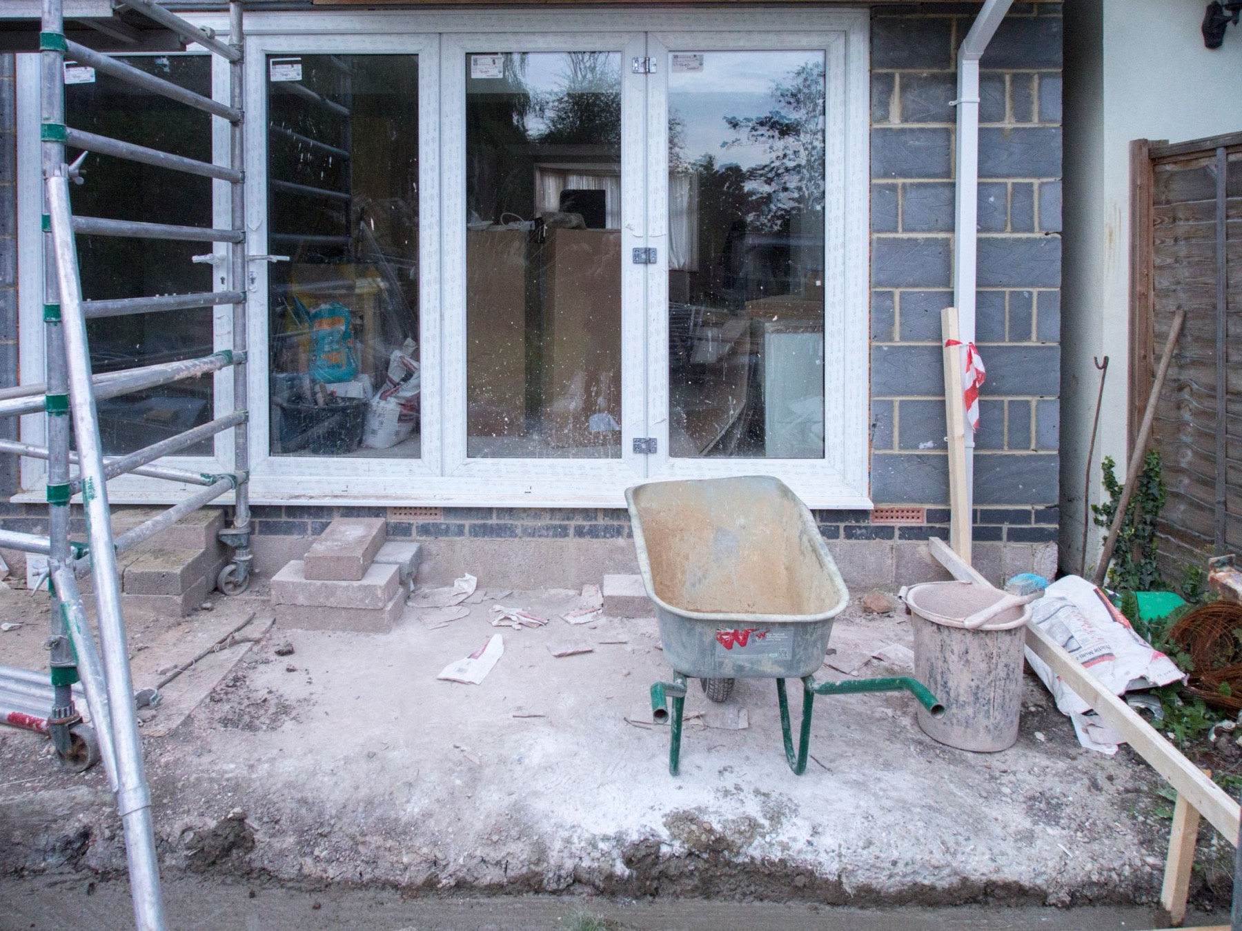 A house part-way through building work — scaffolding up, new blockwork, and a wheelbarrow on the site